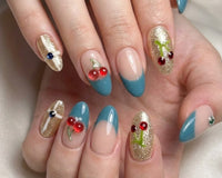 Nude, blue, and gold press-on nails with cherry and rhinestone accents on a hand, combining glossy finishes and detailed nail art for a styl