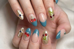 Nude, blue, and gold press-on nails with cherry and rhinestone accents on a hand, combining glossy finishes and detailed nail art for a styl