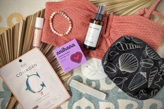 These Palm People box products are displayed on a beach towel: shower steamer, bracelet, small bag, tanner, perfume, and face mask.