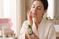 Woman in a white spa robe applying Queen City Skincare Calming Facial Serum to her face beside an open pink skincare box, folded towels, gre