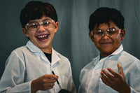 Two children in lab coats and safety glasses are laughing. One is holding a cup and the other is holding a fingerprint brush. 