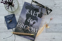 Book title Salt and Broom, glasses, feather bookmark, evil eye necklace, gold pen on a light wood backdrop and a glass bottle with flowers