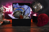open black box sitting in front of a neon light filled with a book, a jar, feathers, stickers and other witchy items