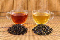 Blacksmith Blend brewed deep amber and Blueberry Lemon Oolong brewed golden in glass cups, each beside its loose leaf on bamboo