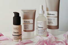 Four brown skin care products from the brand Ordinary. With pink flower leaves layered around the products 