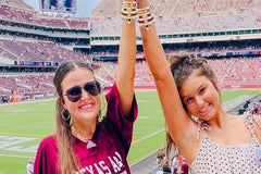 Two women at a football stadium raising their arms and showing off their Game Day stackable friendship bracelet sets with beaded bracelets a