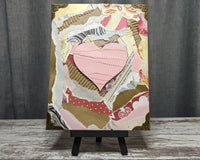 Torn paper art piece on a wooden easel. Layers of pink, red and gold paper are arranged around a pink torn paper heart.
