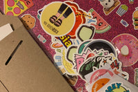 pink glitter binder covered in stickers, overlaid with a stack of stickers ready to be used, and a brown rigid mailer, original packaging