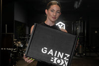 This photo shows an attractive and athletic female model holding a closed Gainz Box in a dark, gritty Los Angeles gym.