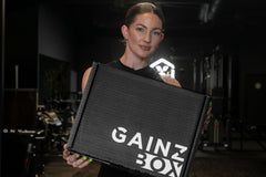 This photo shows an attractive and athletic female model holding a closed Gainz Box in a dark, gritty Los Angeles gym.