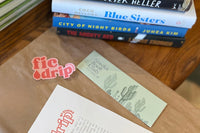 A pile of hardcover books and a smattering of other items including a bookmark a sticker, and a half sheet of paper that says FicDrip.