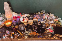 a large quantity of witch related items including dried herbs, crystals, decor and divination tools. lit candles and flowers around them