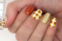 styled nail wraps displaying a bold plaid pattern, golden glitter and a solid burnt orange color, with the thumb featuring a plaid pumpkin