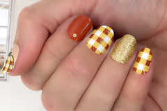 styled nail wraps displaying a bold plaid pattern, golden glitter and a solid burnt orange color, with the thumb featuring a plaid pumpkin