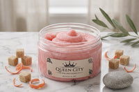 Open jar of pink Queen City Skincare body scrub displayed on marble with sugar cubes, citrus peel, greenery, and a smooth spa stone.