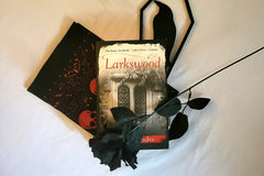 This is a picture of a book titled Larkswood with an artificial black rose, a raven wall decoration, and a skull bandana.