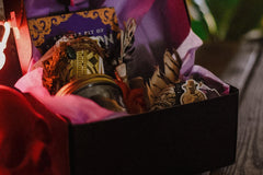 an up close shot of an open box filled with a book, feathers, tarot cards, a jar, a candle and stickers