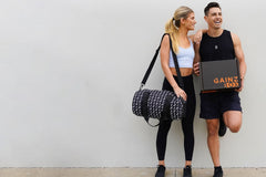 This image shows an attractive female model with a duffel bag standing next to a fit male model holding a closed Gainz Box.