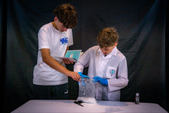 Two children are conducting an experiment at a table. One is holding the instructions while the other is mixing water and plaster in a bag. 