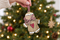 A hand holding the wooden gingerbread man ornament by its gingham ribbon loop in front of a softly lit Christmas tree
