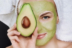 Lady with towel on her head and a half open avocado being held in front of her eye with the avocado mask being displayed on her face