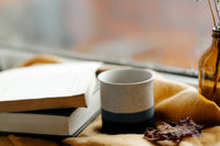 A STACK OF BOOKS SITS BESIDE A STONEWARE COFFEE MUG. A MUSTARD BLANKET IS SPREAD OVER A TABLE. EVERYTHING SITS NEAR A WINDOW.