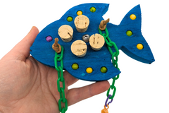 A hand holding a fish shaped piece of balsa wood. It is blue and embedded with cork stoppers and wooden beads. Also has plastic chain. 