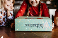 two children are sitting at a wooden dining room table. they are holding a closed wiggle worms kit box. there are digitized snowflakes