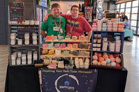 The SoapyFluffs market booth: two people behind a tiered stand of handmade soap bars, with body butters, bath bombs and a banner