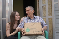 An elderly man receiving a Grandbox gift box alongside a young woman, an emotional scene of care and family connection.
