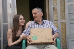 An elderly man receiving a Grandbox gift box alongside a young woman, an emotional scene of care and family connection.