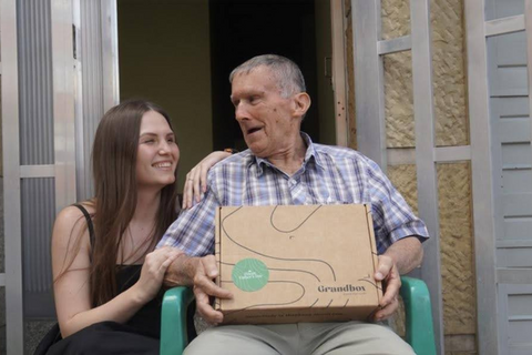 An elderly man receiving a Grandbox gift box alongside a young woman, an emotional scene of care and family connection.