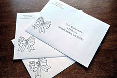 Three white envelopes with printed sample U.S. mailing addresses and black outline patriotic bow designs arranged on a wooden table.