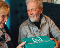 Grandparents smiling together while holding a Senior Joy Box, a monthly subscription gift that brings joy and love.