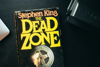 Close up photo of a book called the dead zone from stephen king and a black object in top right corner
