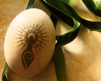This image depicts a wood egg engraved with the Goddess of fertility & emerging sun rays beaming from behind. Green velvet ribbon encircles.