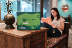A woman reading a book in a cozy nook with a Box of Books subscription box nearby