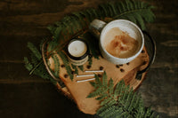 A small glass jar candle is shown sitting on a fern leaf atop a wooden board. Matches lie beside it as well as a cup of coffee.