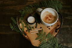 A small glass jar candle is shown sitting on a fern leaf atop a wooden board. Matches lie beside it as well as a cup of coffee.