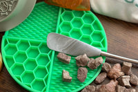 Image of a green lick mat on a table. On top of the green lick mat are freeze dried dog treats and a peanut butters spreader.