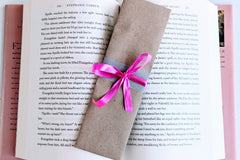 A Romance-themed bookmark is wrapped in brown craft paper and decorated with pink ribbon. It rests on an open hardback book.