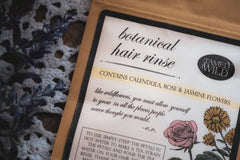 kraft bag with large label titled botanical hair rinse with an ingredient list all next to a purple sprig on top of a white doily