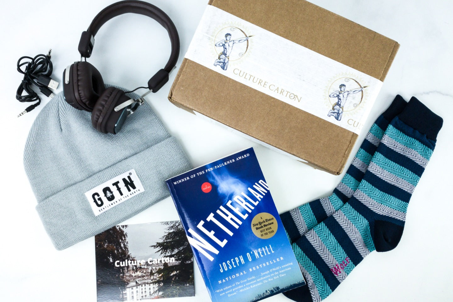 A subscription box labeled Culture Carton surrounded by headphones, a knit hat, striped socks, and a book titled Netherland.