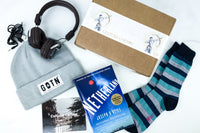 A subscription box labeled Culture Carton surrounded by headphones, a knit hat, striped socks, and a book titled Netherland.
