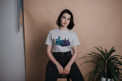 A young woman sitting on a stool wearing a Harry Potter Hogwart's Castle Tshirt in greys, purples, blues, and greens.