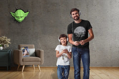 A man wearing a Death Star Tshirt and young child play videos games. There is a 3D Yoda light hanging on the wall and a Journal in a chair.