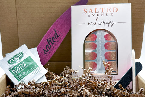 Box with Nail Wraps, bracelet, nail file, alcohol prep pads, mini nail file and a cuticle tool resting in a box filled with crinkle paper.