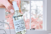 Hand holding the Soothe + Sleep Lavender Bath Oil by Greenmade goods over an antique bath tub with pink flowers in the background.
