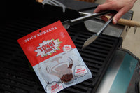 A bag of Tong Spicy Sriracha jerky lying on gas grill grates with a man's hand holding grilling tongs picking up the bag. 
