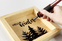 A hand holding a woodburning tool that is burning the words "Hello Winter" over the top of various pine trees.